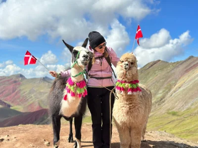TourCompass-Mitarbeiterin Iida steht auf dem Gipfel des Rainbow Mountain in Peru und hält zwei Flaggen sowie ein Alpaka in den Händen.