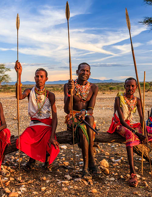 Eine Gruppe Massai sitzt in der Savanne, Speere in den Händen.